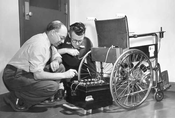Two men looking at the battery of the George Klein Power Wheelchair   